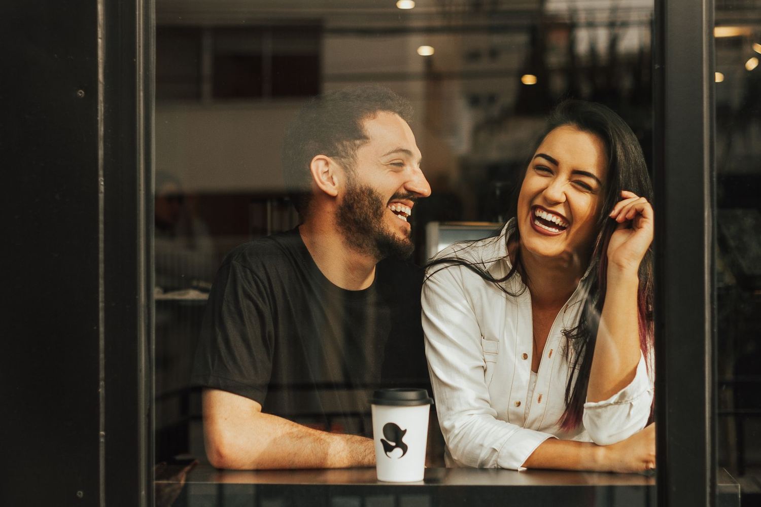 Two people laughing at a table behind a cafe window